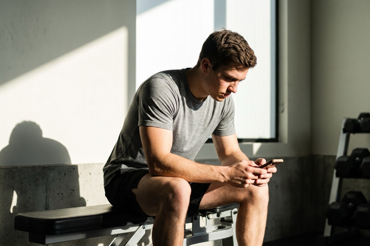 Man sitting on a gym bench between sets, head down, looking at his phone