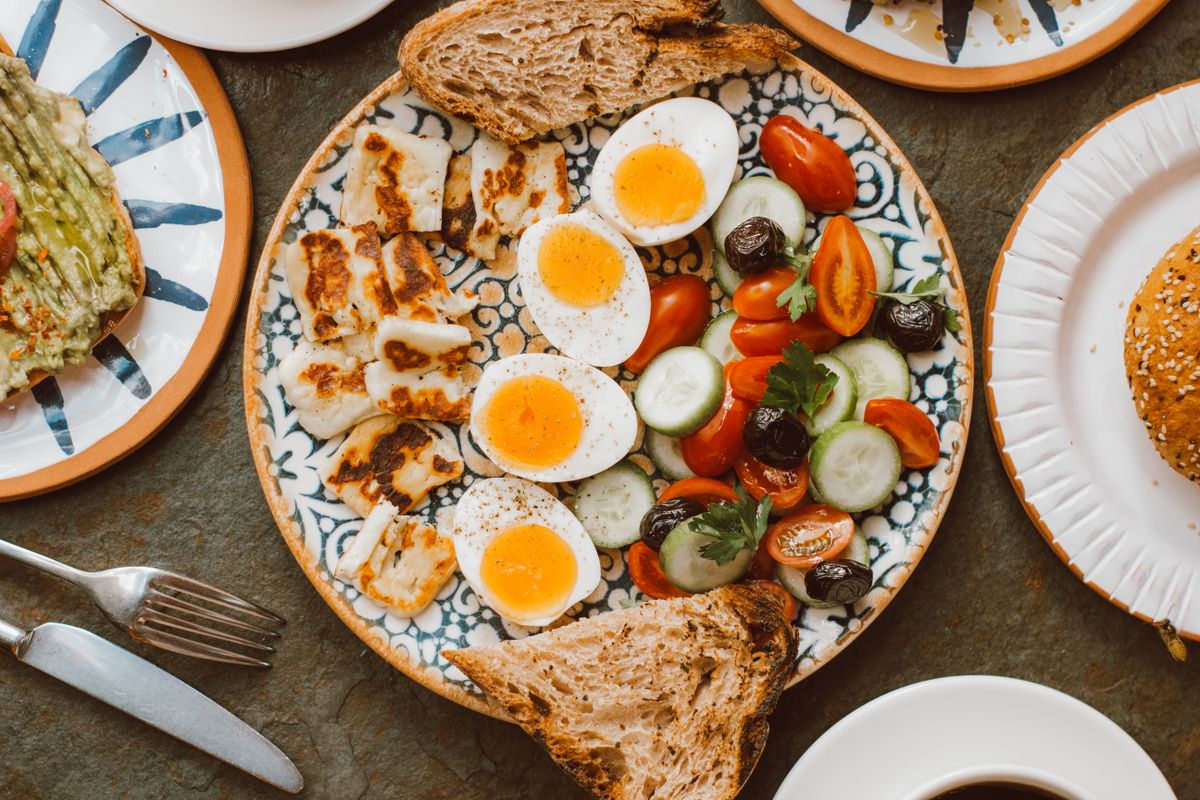 Overhead view of plates of boiled eggs, grilled halloumi, tomatoes, cucumber, olives and bread