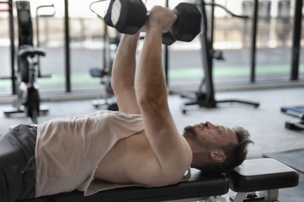 Grey-haired man pressing a pair of dumbbells overhead on a gym bench
