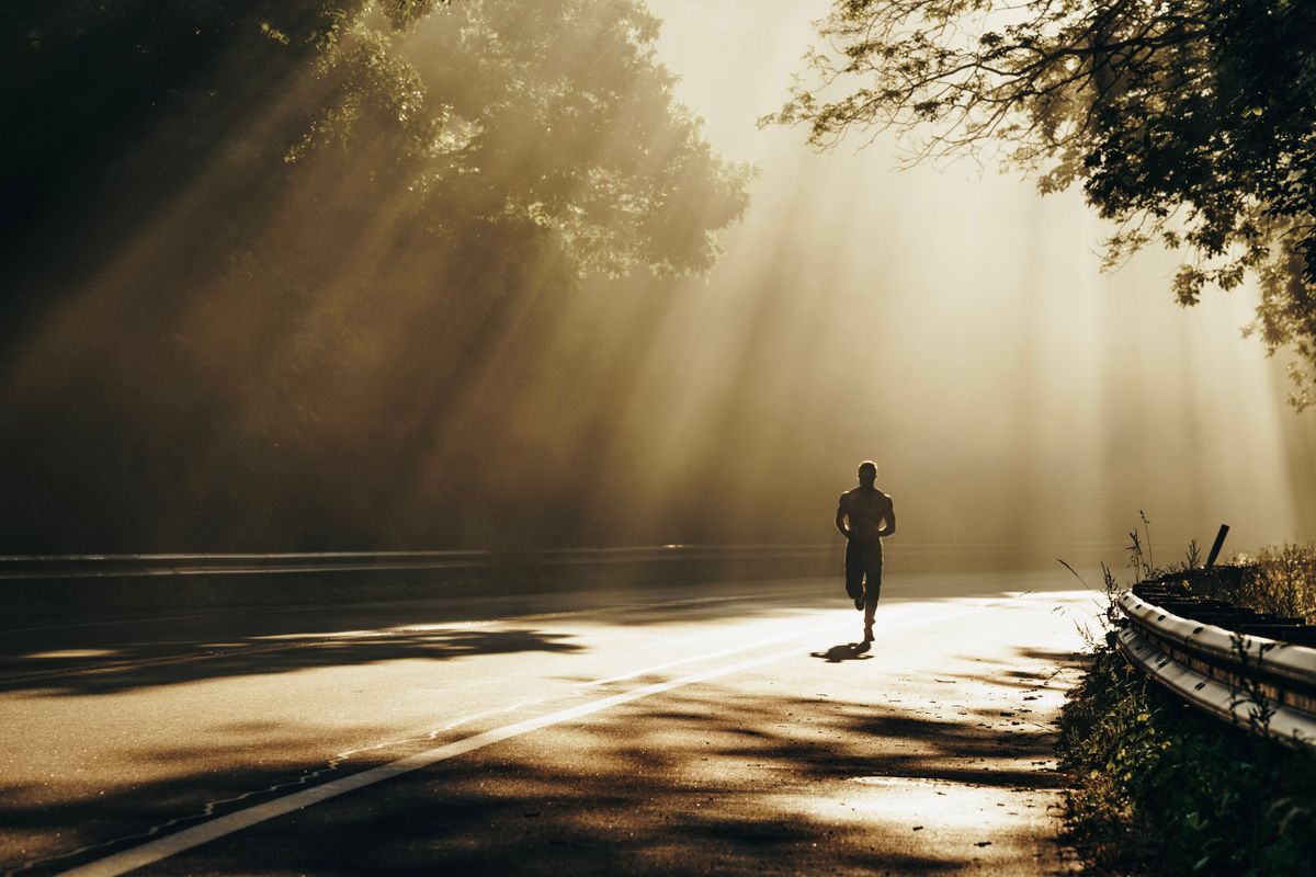 Lone runner silhouetted on a forest road as low sun beams through the trees