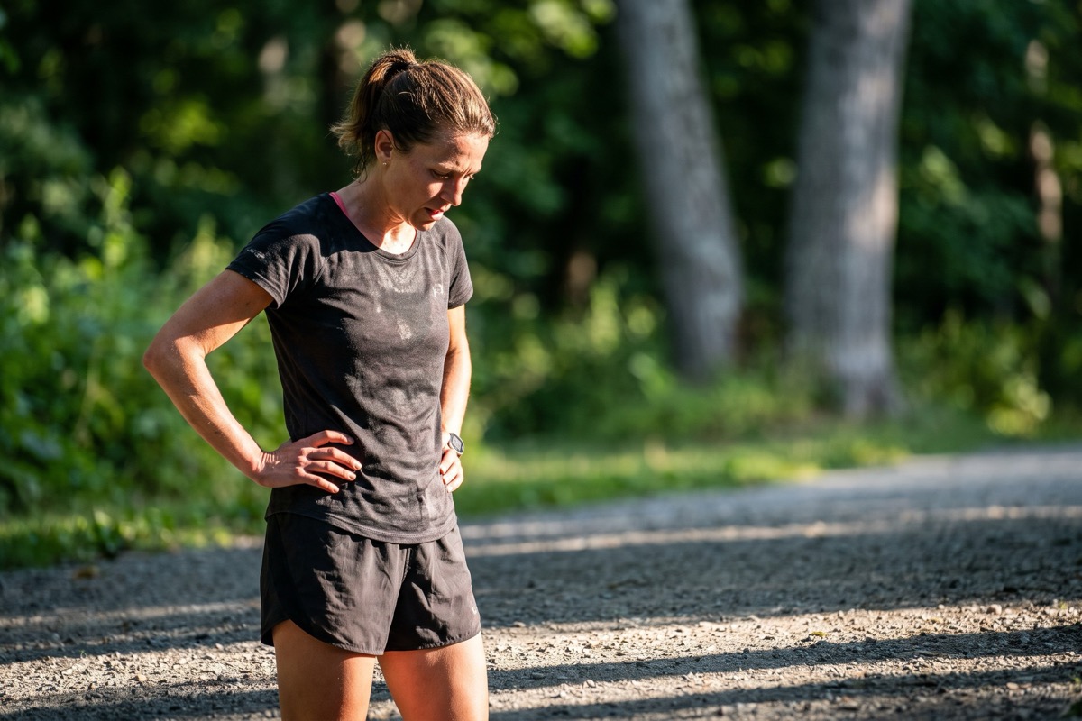 A runner stopped to walk on a park path, hands on hips, catching her breath